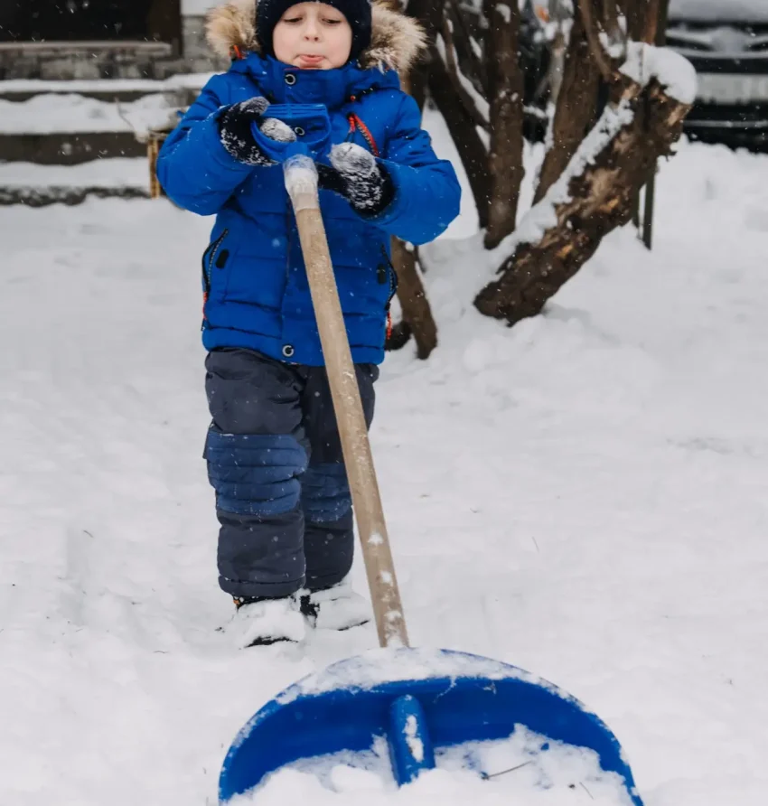 Little boy with a shovel in hand remove snow in backyard, snow removal. Kid in blue jacket cleans